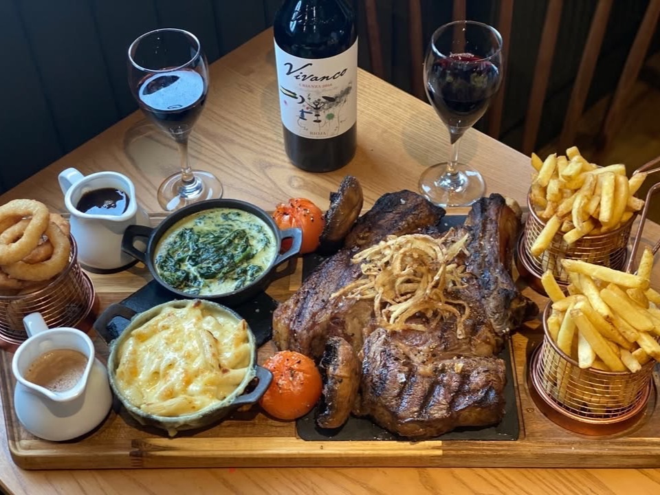 Two steaks on a board with chips, sides and a bottle of red wine